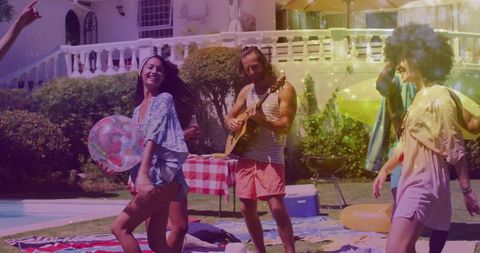 Friends dancing by pool while man plays guitar at sunlit backyard summer party with floats