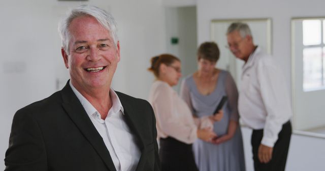 Senior Businessman Smiling at Camera with Colleagues in Background - Download Free Stock Images Pikwizard.com