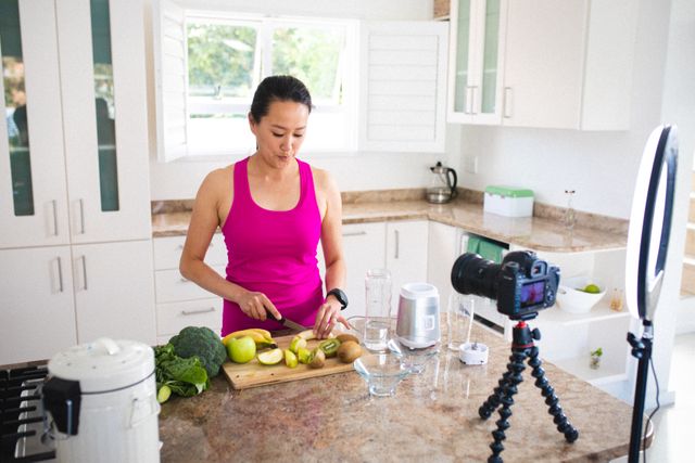 Asian woman preparing healthy juice by cutting fruits in a modern kitchen while vlogging. Ideal for content related to healthy lifestyle, cooking vlogs, influencer marketing, and technology in communication. Useful for websites, blogs, and social media posts promoting healthy eating, home cooking, and digital content creation.