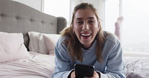 Smiling woman lying on bed holding smartphone wearing hoodie and socks, cozy pastel bedroom lifestyl