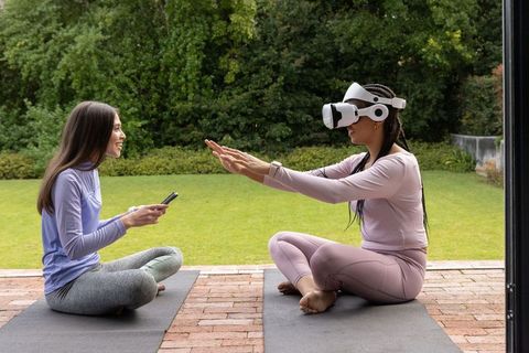 Women using vr headset and smartphone while doing yoga on backyard mats for home wellness
