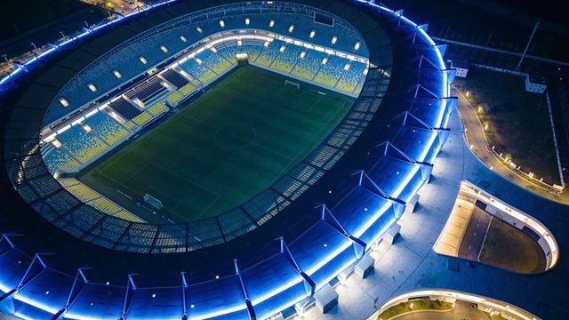Aerial nighttime view of illuminated oval stadium with blue led ring and empty grass pitch