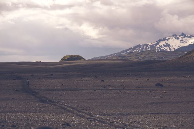 Wide view of a stark desert with rugged rocky terrain leading up to a snow-capped mountain in the background. Cloudy sky adds dramatic effect. Ideal for use in nature, adventure, and travel content to depict rugged, remote beauty. Suitable for themes around exploration, isolation, or tranquility.