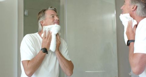 Mature man drying face with towel at bathroom mirror home grooming routine