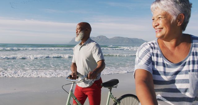 Senior couple enjoying a walk on the beach while pushing their bicycles. Man dressed in a white polo and red trousers, woman in a striped shirt. Background features blue ocean waves and mountain scenery. Image suitable for promoting healthy lifestyles, retirement communities, medical services, vacation destinations, and outdoor activities.