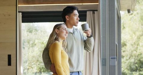 Modern couple embracing by sunlit sliding door holding mug overlooking lush green backyard