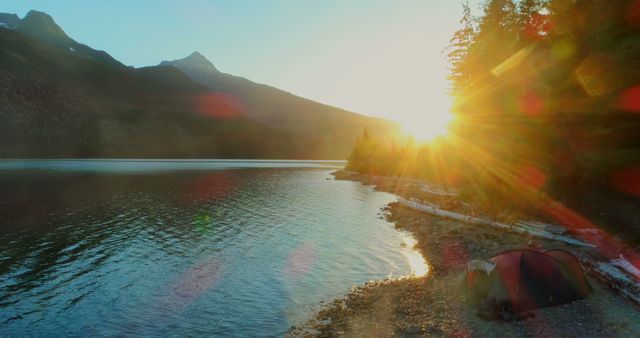 Sun rising over pristine lake with tent set up on wooded shore, mountains in background. Ideal for travel blogs, outdoor adventure websites, nature magazines, and promoting camping gear.