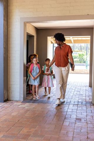 Early childhood teacher walking with young students guiding through sunlit brick corridor