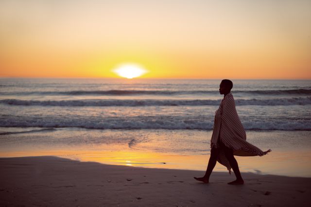 This image captures a woman wrapped in a blanket walking along the beach during sunset. The serene and peaceful setting with the ocean waves and the horizon in the background makes it perfect for themes related to relaxation, tranquility, nature, and solitude. It can be used in travel blogs, wellness websites, inspirational content, and advertisements promoting beach destinations or relaxation products.