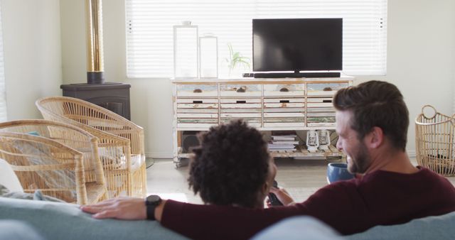 Image of back view of happy diverse couple drinking coffee and watching tv with copy space. Love, relationship and spending quality time together at home.