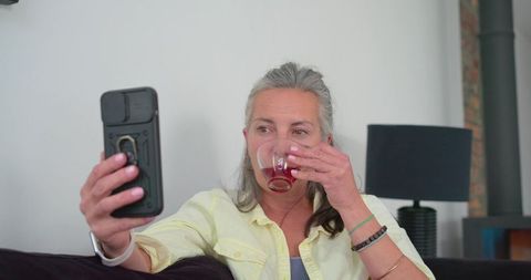 Mature woman taking selfie while sipping red drink on sofa in cozy living room