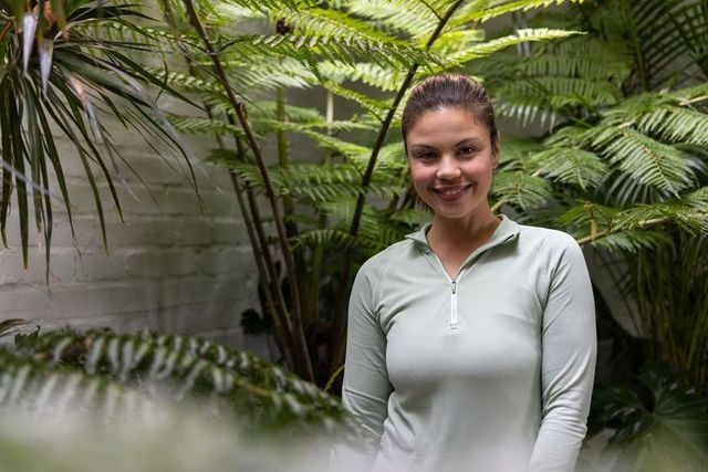 Asian woman smiling among lush ferns in greenhouse wearing light green zip-top, relaxed