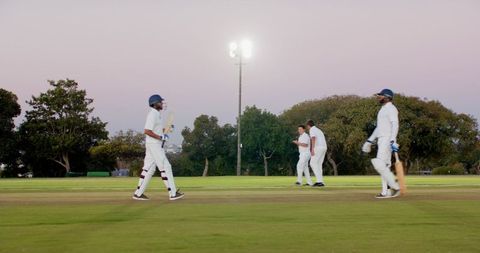 Cricket players walking at dusk on pitch under floodlights during twilight