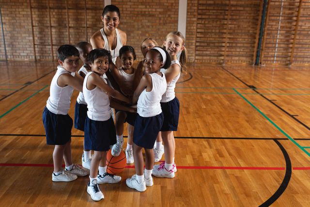 Front view of schoolkids and female coach forming hand stack and looking at camera at basketball court in school