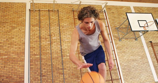 Young man focused and practicing basketball dribbling in a gym. Ideal for use in fitness, sports, exercise content or basketball training programs and ads for sports equipment.