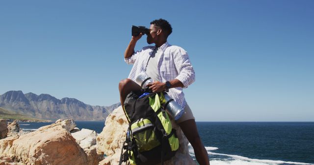 African american man hiking using binocilars sitting on rock by the coast. fitness training and healthy outdoor lifestyle.