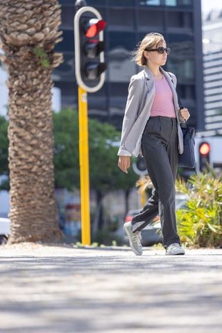 Young woman walking across urban crosswalk wearing light grey blazer and sunglasses
