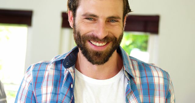 This image shows a smiling bearded man wearing a plaid shirt indoors. Suitable for lifestyle, casual clothing promotions, articles about men's fashion, everyday life, or advertising friendly and approachable personalities.