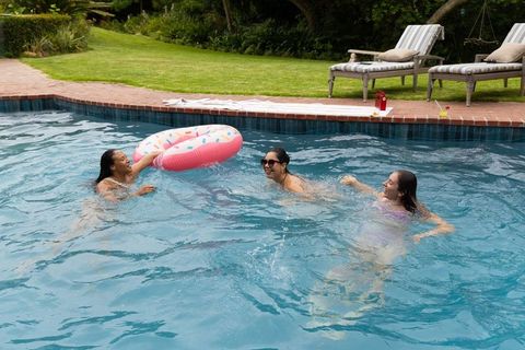 Diverse friends splashing in backyard pool reaching for pink donut float and laughing