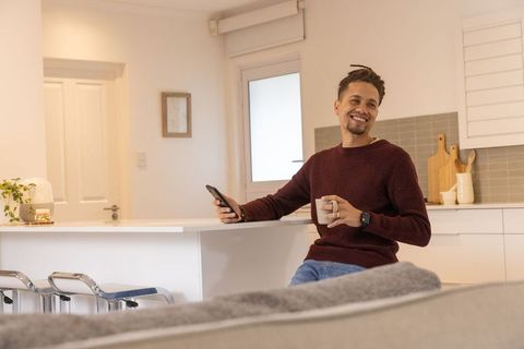 Smiling man relaxing on kitchen island holding smartphone and coffee mug in cozy kitchen