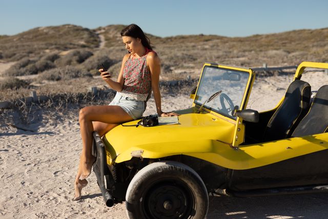 Caucasian woman sitting on beach buggy with camera and map, using smartphone on sunny beach. beach stop off on summer holiday road trip.