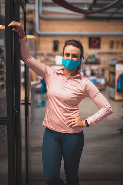 Caucasian woman standing in the doorway of a climbing gym, wearing a face mask and casual activewear. Ideal for illustrating fitness and leisure activities during the COVID-19 pandemic, health and safety measures, or retail environments. Useful for articles, advertisements, and promotional materials related to fitness, health, and pandemic precautions.