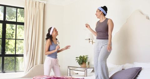 Diverse women enjoying pajama party and morning champagne toast on bed in bright cozy bedroom