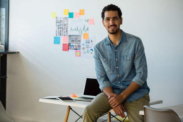 Young man sitting on desk in a modern, creative office, smiling confidently. Ideal for use in business, entrepreneurship, and startup-related content. Perfect for illustrating modern work environments, professional confidence, and casual office settings.