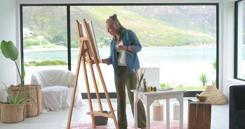 Woman painting at easel in bright waterfront home studio wearing headphones and denim shirt