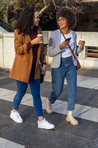 Friends walking and laughing while holding coffee and checking phone on urban sidewalk