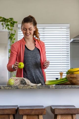 Woman packing reusable mesh bag with fresh produce wearing coral jacket at kitchen counter