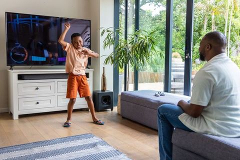 African american father watching son dancing during living room gaming in sunlight