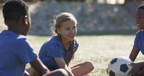Diverse teen soccer teammates sitting on sunny field smiling while holding soccer ball