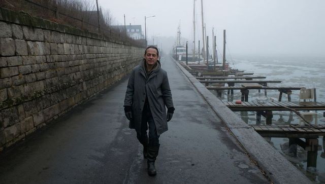 Man walking along foggy harbor quay wearing long gray coat and rubber boots, moored boats