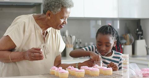 Grandmother guiding granddaughter decorating pink cupcakes in modern home kitchen