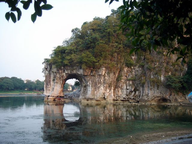 Elephant Trunk Hill is reflected in calm river water, set against a serene sunset in Guilin, China. Ideal for travel websites, nature publications, and promoting tourism in China. This peaceful and scenic view highlights the unique rock formation widely recognized for its resemblance to an elephant trunk dipping into the river.