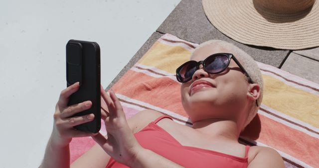 Biracial woman with sunglasses sunbathing lying on towel using smartphone. Lifestyle, free time, summer, communication and vacation, unaltered.