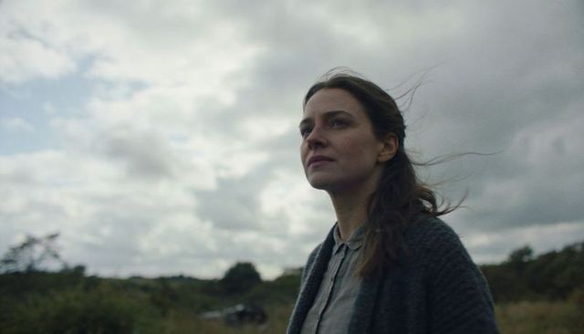 Woman standing in windswept moorland under overcast sky, contemplative cinematic portrait