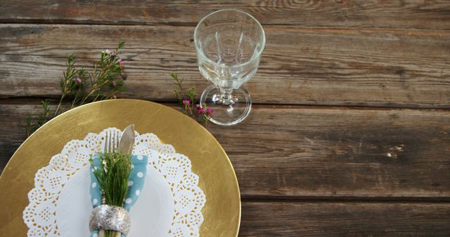 A rustic table setting features a golden charger plate and an elegant glass, with copy space. The arrangement includes silverware wrapped in a polka-dotted napkin, accented by a sprig of flowers and a decorative element, creating a charming and inviting atmosphere.