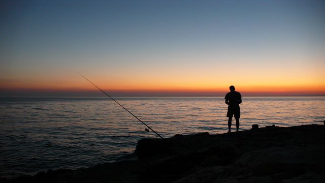 Silhouette of man fishing at sunset on seashore with calm ocean and colorful sky. Ideal for articles, advertisements, or social media posts related to outdoor activities, hobbies, and relaxation by the sea. Perfect for peaceful and reflective themes in travel and leisure content.
