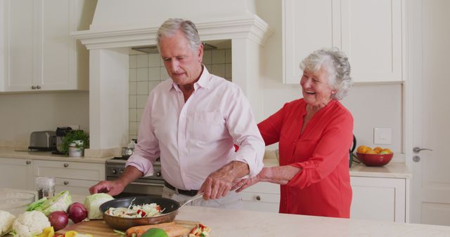 Elderly couple enjoying their time cooking together in a bright modern kitchen. The man is stirring vegetables in a saucepan, while the woman lovingly embraces him from behind, laughing. Ideal for themes of family, love, happiness, healthy living, and togetherness among older adults. Could be used in advertisements, blog posts, or articles related to healthy lifestyles, senior living, and relationships.