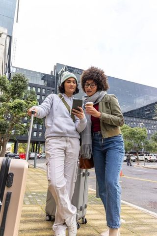 Two friends standing in city plaza checking phone while holding coffee and suitcases
