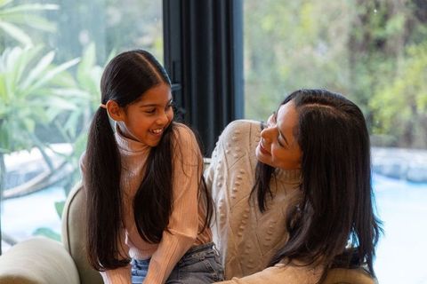 Indian mother and daughter sharing warm smiles on sofa by window in cozy sweaters