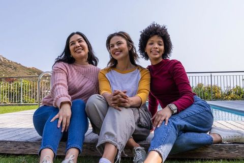 Smiling diverse women relaxing on backyard pool deck wearing sneakers and casual clothes