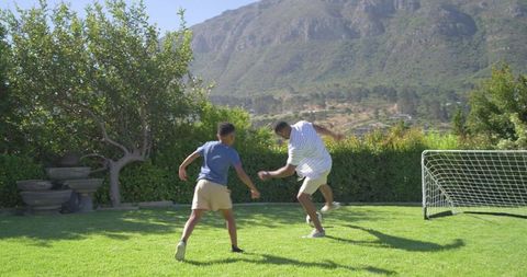 African american father and teenage son playing soccer on sunny backyard lawn