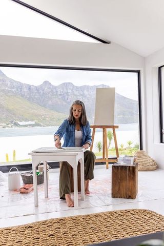 Female artist painting at sunlit lakeside studio with mountain view and easel, bright light