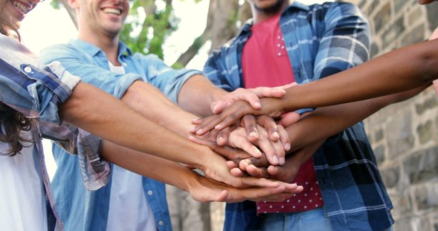 A diverse group of young adults stack their hands together in a gesture of unity and teamwork, with copy space. Their smiling faces and the outdoor setting suggest a casual, friendly atmosphere and the importance of collaboration.