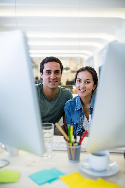 Two young graphic designers are smiling while working together in a modern office. They are sitting at a desk with computers, showcasing a collaborative and creative work environment. This image can be used for promoting teamwork, modern office culture, creative professions, and digital workspaces.