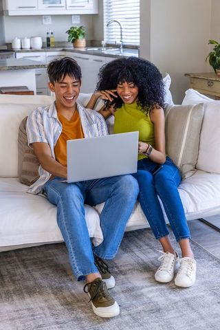 Diverse young couple relaxing on sofa using laptop for remote work and online shopping