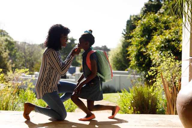 African-American mother kneeling to fix her daughter's facemask before school. Daughter wearing a backpack, ready for school. Scene takes place outdoors in front of their house. Ideal for use in family, education, safety, and health-related content.